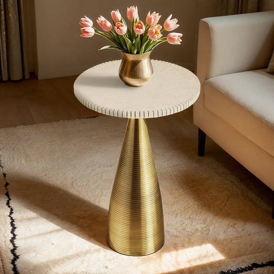 A sunlit, high-rise apartment living room featuring the Oasis End Table with a notched sandstone top and a gold conical base, styled with a vase of pink tulips next to a beige sofa.